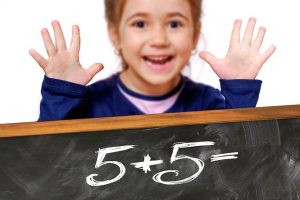 Na pierwszym planie działanie napisane na tablicy 5+5=, w tle dziewczynka pokazująca 10 paluszków